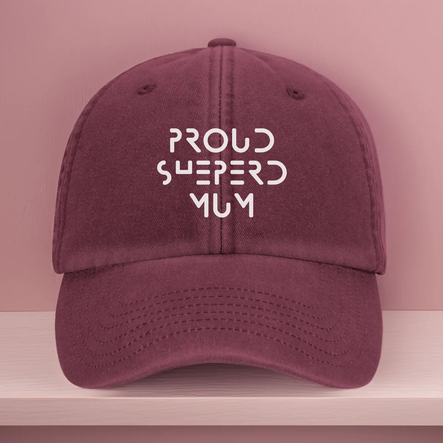 Vintage-style burgundy baseball cap with white embroidery that reads "Proud Shepherd Mum" in a bold modern font. Designed as a personalised gift for German Shepherd dog mums and proud dog parents.
