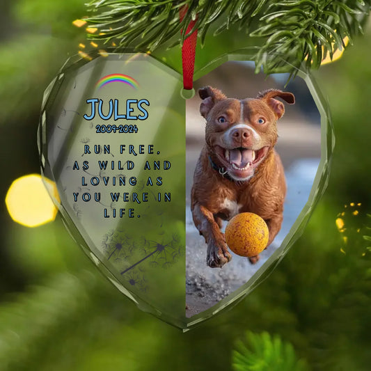 Heart-shaped glass ornament hanging on a Christmas tree, featuring a joyful brown dog running on the beach with a ball. The left side displays the name “Jules,” the dates “2009–2024,” a rainbow, and the tribute “Run free. As wild and loving as you were in life.”