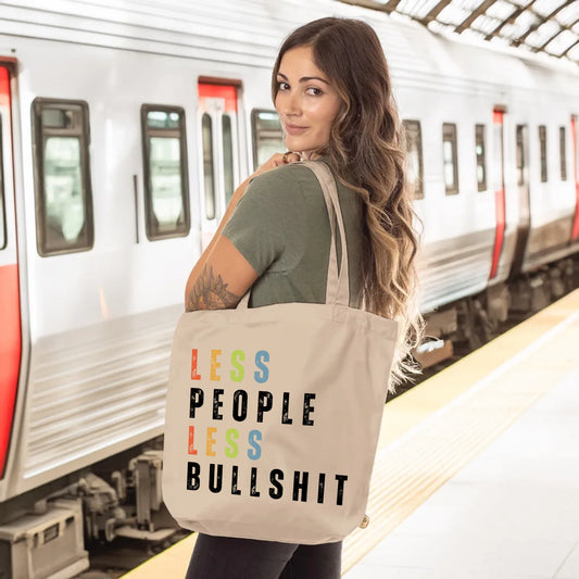 Woman standing on a train platform with a neutral-toned tote bag that reads “LESS PEOPLE LESS BULLSHIT” in colorful, bold letters. Ideal for dog lovers who foster and value peace over chaos. The back says, “that’s why I foster dogs,” making it perfect for proud dog foster carers with a sense of humor.
