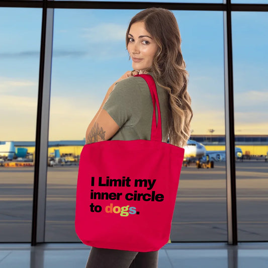 Bright red organic tote bag with the phrase “I limit my inner circle to dogs.” in bold black and rainbow text, held by a woman in an airport setting. The front side says: “No people. No bullshit.”